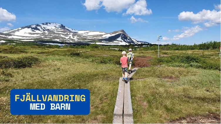 Fjällvandring med barn, två barn syns vandra på spängerna på väg mot en skylt längs vandringsleden. I bakgrunden syns ett stort fjäll med en snötäckt topp. Solen skiner.