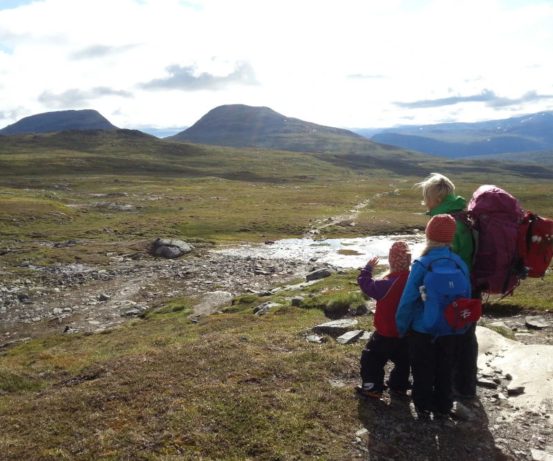 foto från fjällvandring barnfamilj där en förälder ses bära egen ryggsäck och minsta barnets ryggsäck fäst baktill, det äldre barnet bär sin egen ryggsäck, familjen blickar ut över fjällen från Blåhammaren ner mot Storulvån